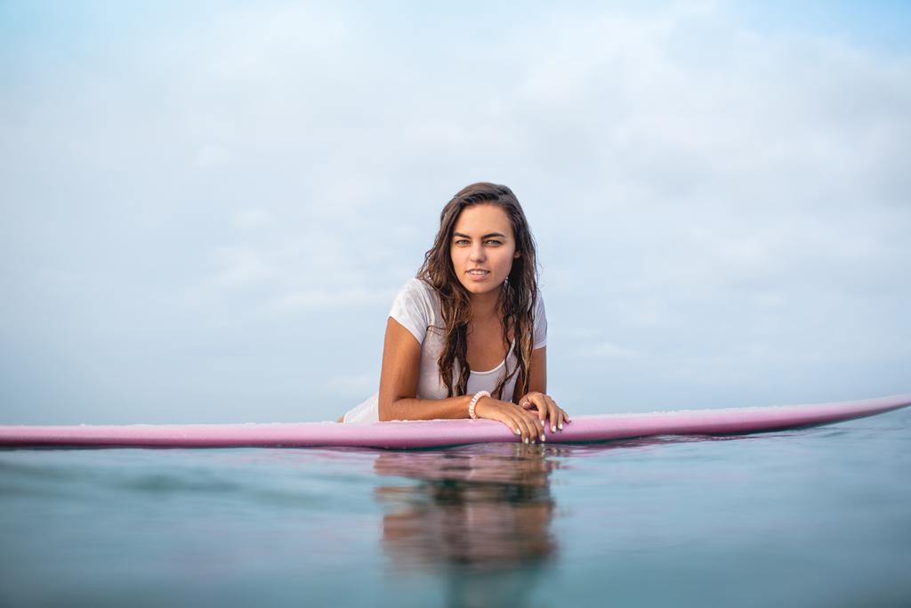 attraktive junge Frau blickt auf Surfbrett im Meer in die Kamera - Foto, Bild