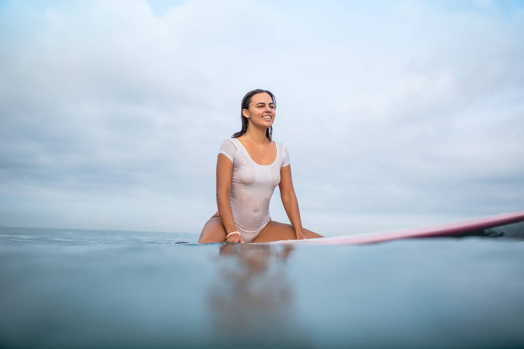 smiling attractive woman in white wet swimsuit sitting on surfboard in ocean during summer vacation - Photo, Image