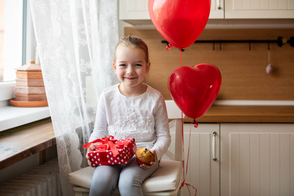 Şirin iskambil kız 6 doğum günü kutluyor. Onun doğum günü kek ve balonlar tarafından çevrili mutfakta oturmuş güzel sarılmış hediye tutan kız. - Fotoğraf, Görsel