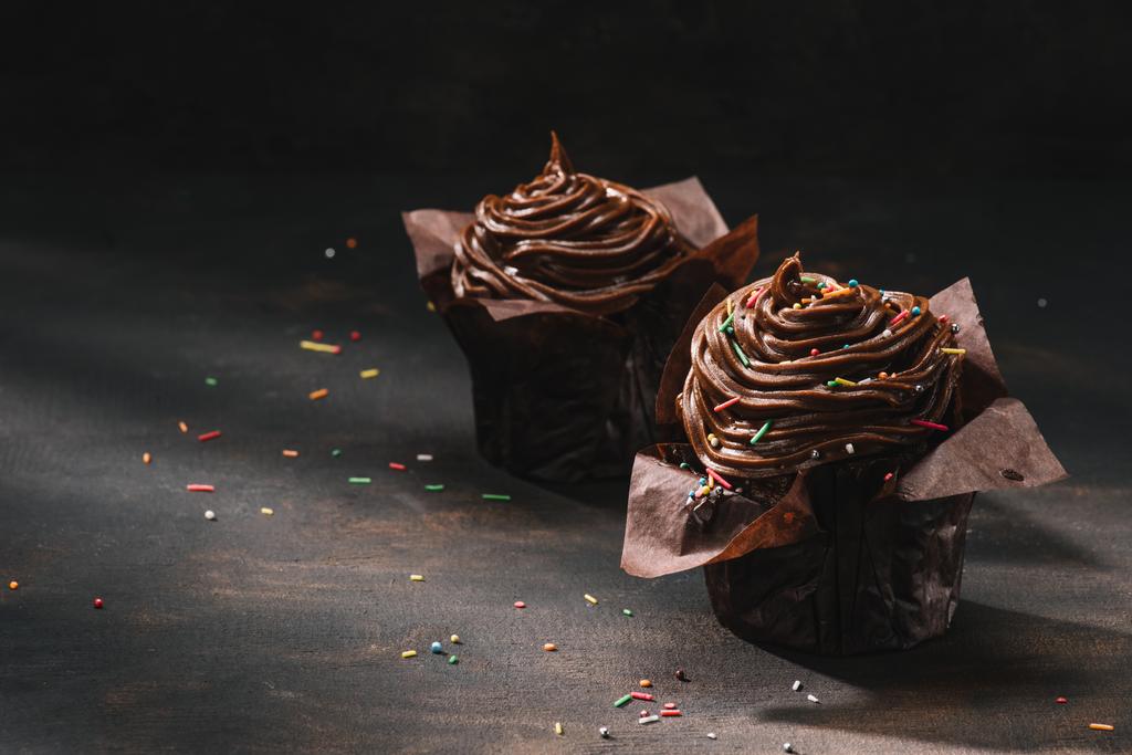 Яскраві шоколадні кекси з масляною глазур'ю та цукром, що розкидані на столі
 - Фото, зображення