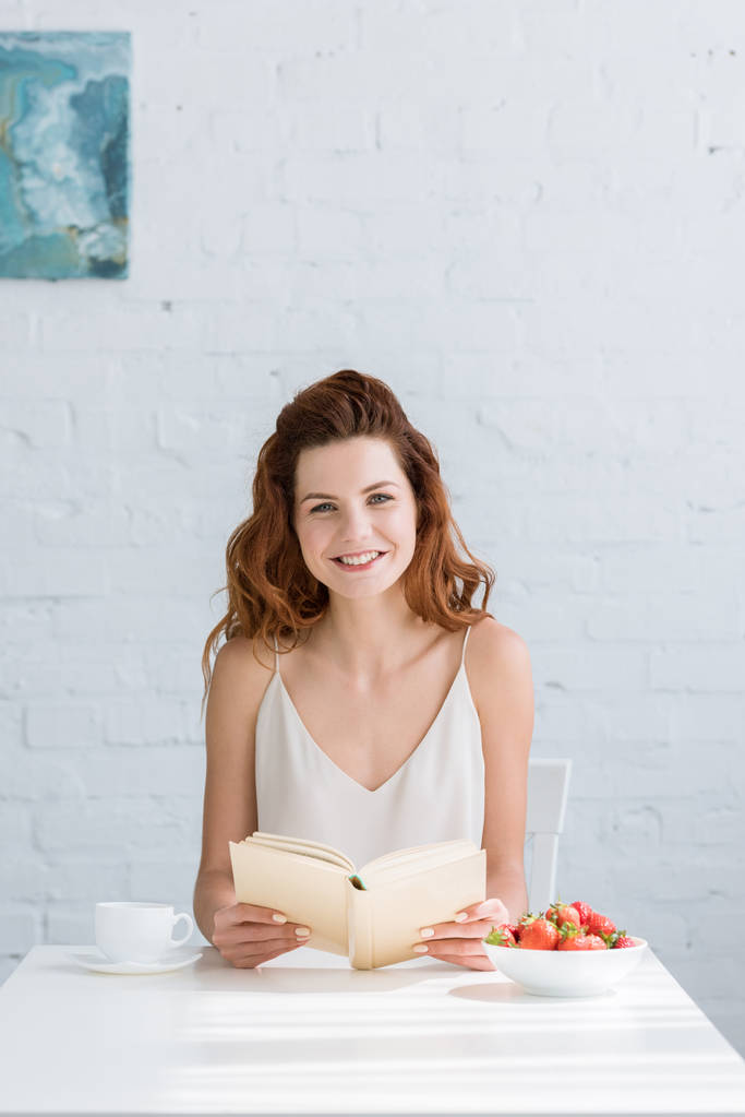 glimlachend jonge vrouw zittend aan tafel met koffie en aardbei en lezen boek thuis - Foto, afbeelding