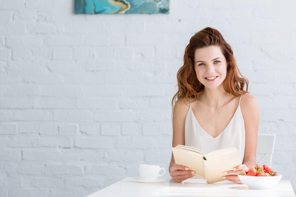 glückliche junge Frau sitzt bei Kaffee und Erdbeere am Tisch und liest zu Hause Buch - Foto, Bild