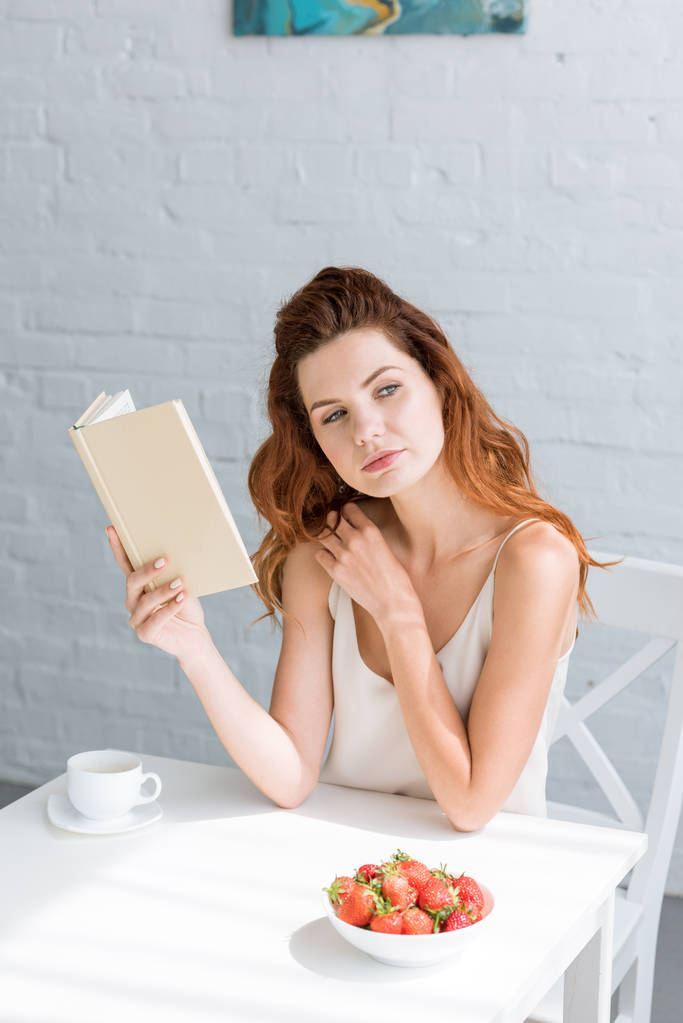 усвідомлена молода жінка з книгою і чашкою кави, сидячи за столом
 - Фото, зображення