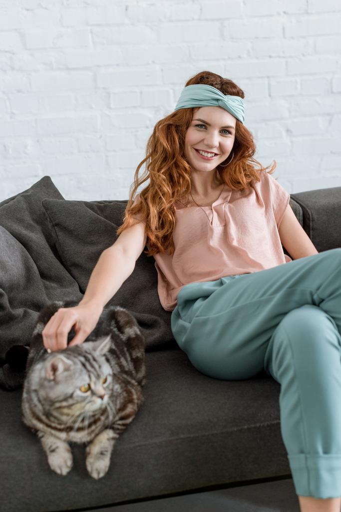 sonriente joven mujer acariciando tabby gato mientras sentado en sofá en casa
 - Foto, imagen