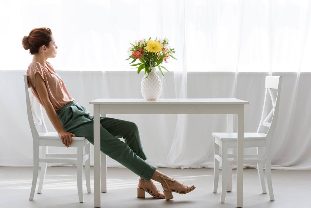 вид сбоку привлекательной молодой женщины, сидящей дома за столом с красивыми цветами в вазе
 - Фото, изображение