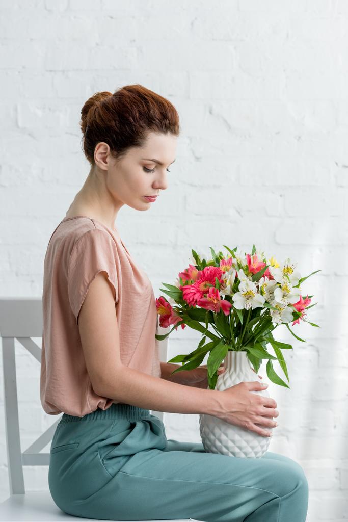 Zijaanzicht van aantrekkelijke jonge vrouw vaas met bloemen te houden terwijl zittend op een stoel voor witte bakstenen muur - Foto, afbeelding
