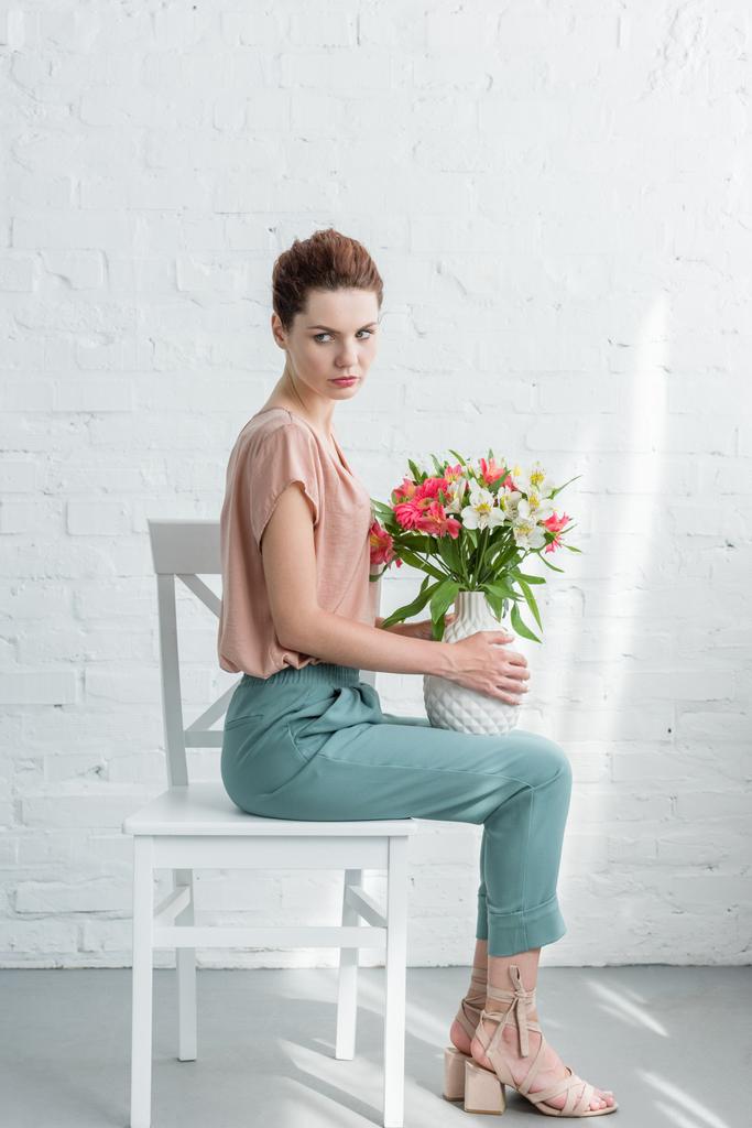 vista lateral de la hermosa mujer joven sosteniendo jarrón con flores mientras está sentado en la silla delante de la pared de ladrillo blanco
 - Foto, imagen