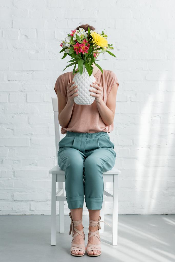женщина покрывает лицо цветами в вазе, сидя на стуле перед стеной из белого кирпича
 - Фото, изображение