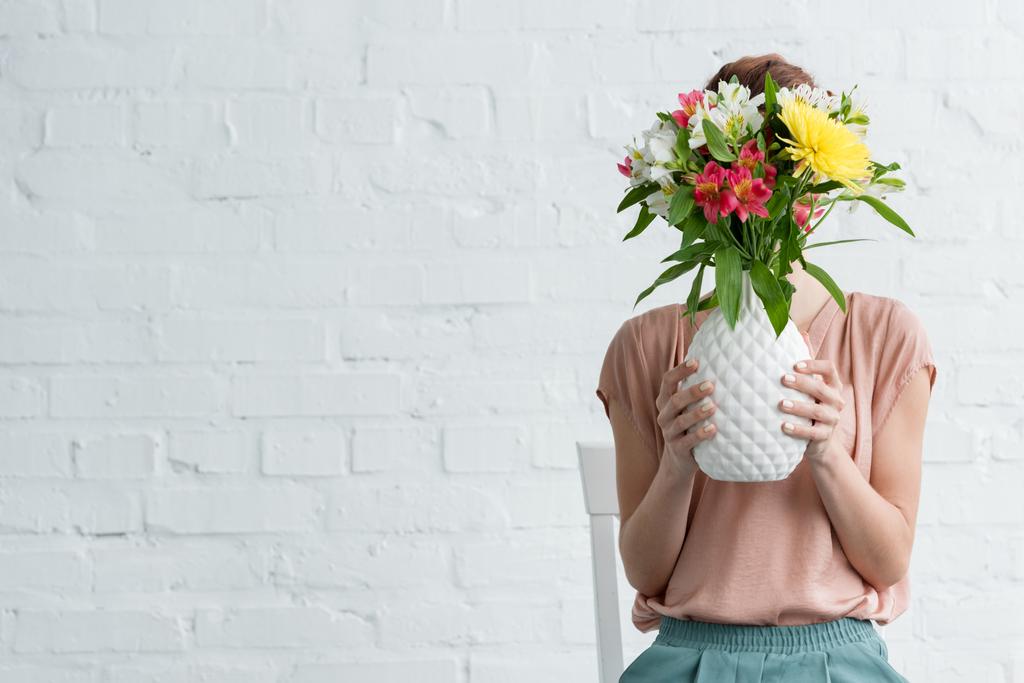 vrouw die betrekking hebben op gezicht met bloemen in vaas voor witte bakstenen muur - Foto, afbeelding