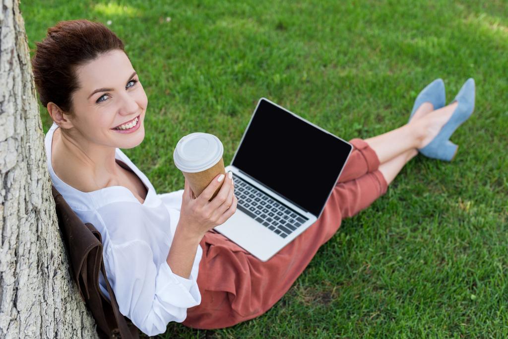 visão de alto ângulo de feliz jovem mulher trabalhando com laptop enquanto sentado na grama com xícara de papel de café e olhando para a câmera
 - Foto, Imagem