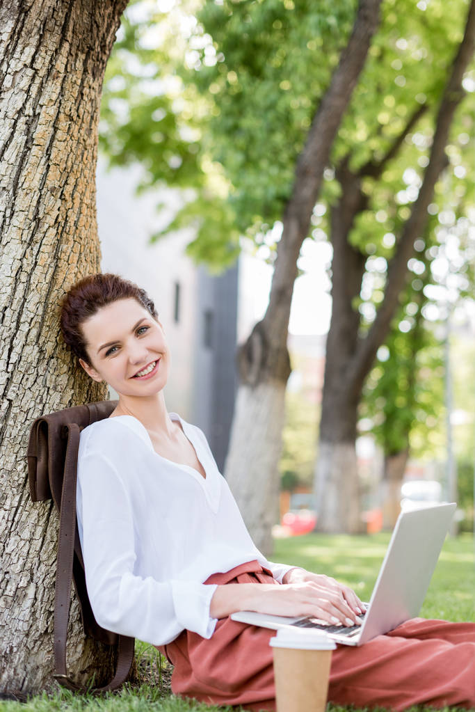 glückliche junge Freiberuflerin arbeitet mit Laptop, lehnt sich im Park auf Baumstamm zurück und blickt in die Kamera - Foto, Bild