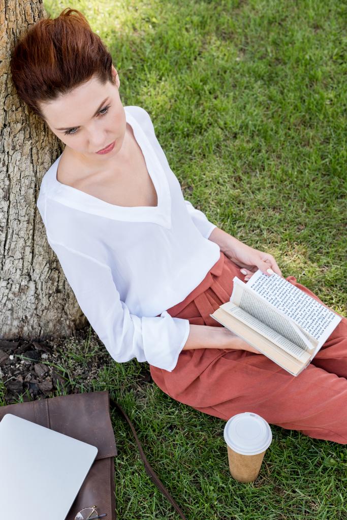 high angle view of beautiful young woman with book leaning back on tree trunk in park - Photo, Image