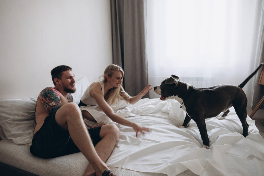 Getatoeëerde paar zittend op bed en spelen met de hond thuis - Foto, afbeelding