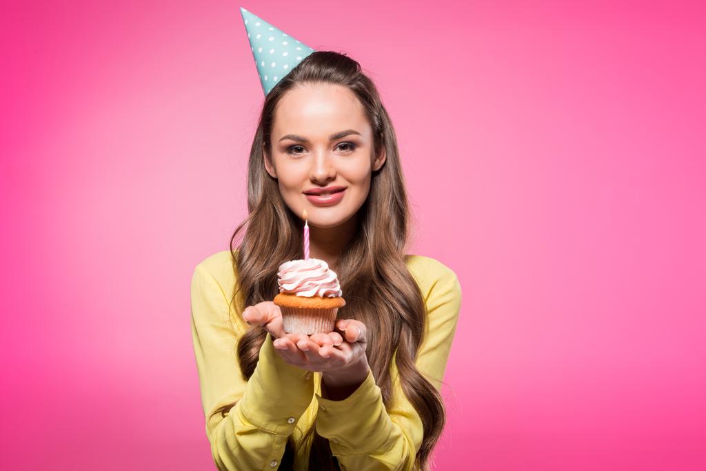 attraktive Frau mit Partyhut, Cupcake in der Hand und Blick in die Kamera isoliert auf rosa - Foto, Bild
