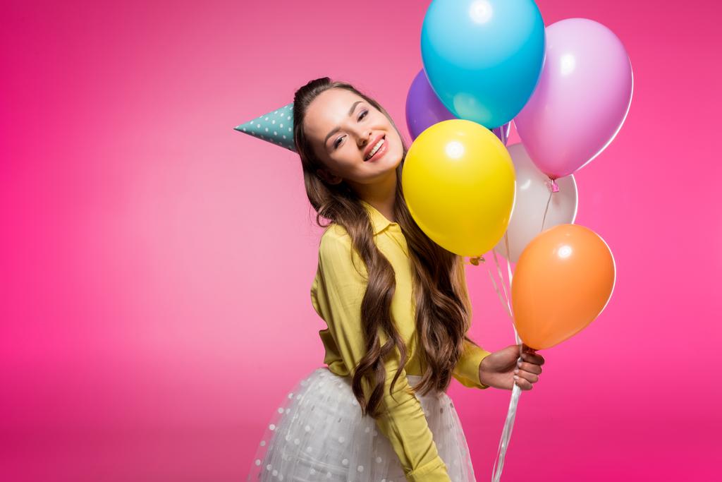 ελκυστική γυναίκα που παρουσιάζουν με καπέλο του πάρτυ και μπαλόνια που απομονώνονται σε ροζ - Φωτογραφία, εικόνα