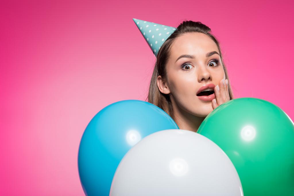 потрясенная привлекательная женщина в шляпе вечеринки и воздушных шарах изолированы на розовый
 - Фото, изображение