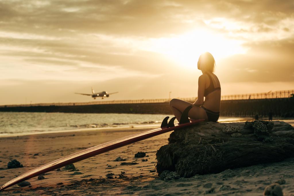 woman sitting on rock with surfboard on beach at sunset with airplane in sky - Photo, Image