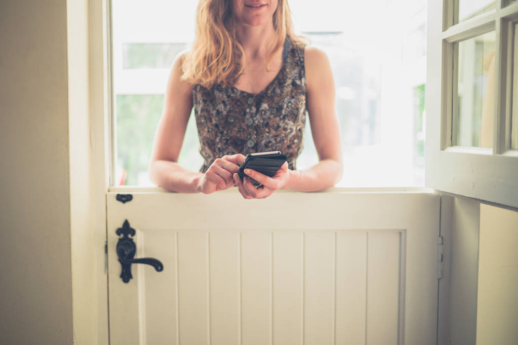 Молода жінка стоїть біля стабільних дверей і використовує свій смартфон
 - Фото, зображення