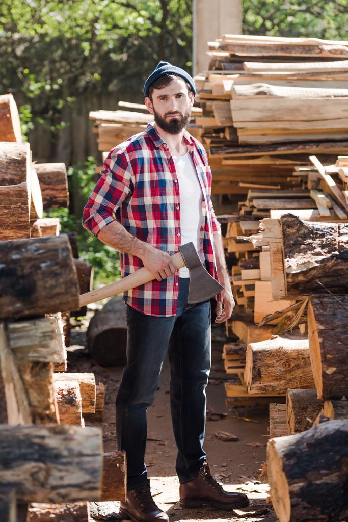 ξυλοκόπος σε καρώ πουκάμισο στέκεται με τσεκούρι μεταξύ κορμών στο πριονιστήριο  - Φωτογραφία, εικόνα