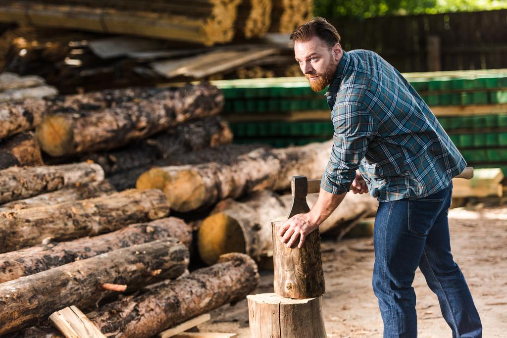 bearded lumberjack in checkered shirt chopping log at sawmill  - Photo, Image