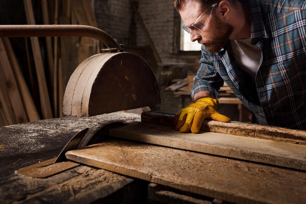 Schnappschuss eines bärtigen Zimmermanns in Schutzhandschuhen und Brille mit der Motorsäge im Sägewerk - Foto, Bild