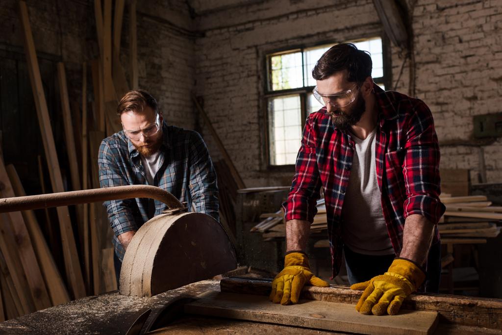 Brodaty carpenter w gogli ochronnych i rękawic przy użyciu piły maszyny i partnera, stojąc w pobliżu Tartak - Zdjęcie, obraz