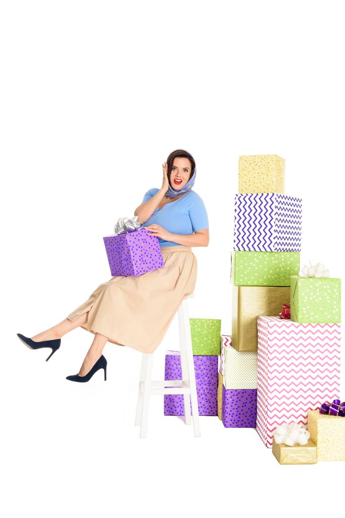 έκπληκτος pin γυναίκα κρατώντας κιβώτιο δώρων και να κάθεται σε σκαμνί κοντά σε σωρούς από δώρα που απομονώνονται σε λευκό   - Φωτογραφία, εικόνα