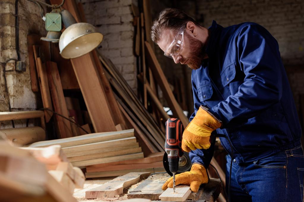 Brodaty carpenter w rękawice ochronne i googles za pomocą wiertarki elektryczne w tartaku - Zdjęcie, obraz
