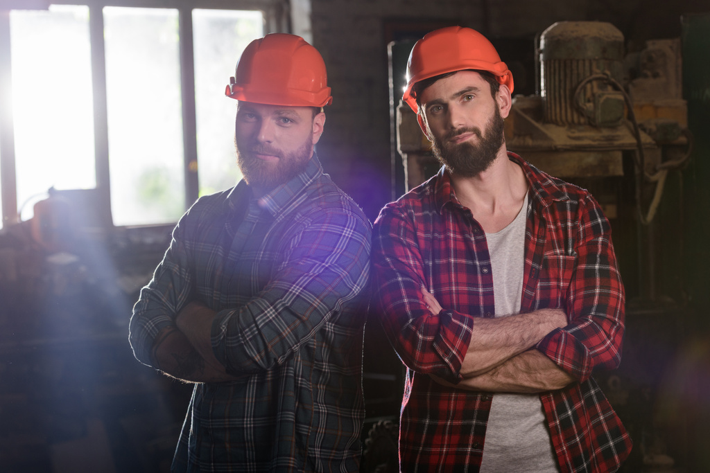 dois carpinteiros barbudos com as mãos cruzadas em capacetes de proteção em pé na serraria
  - Foto, Imagem