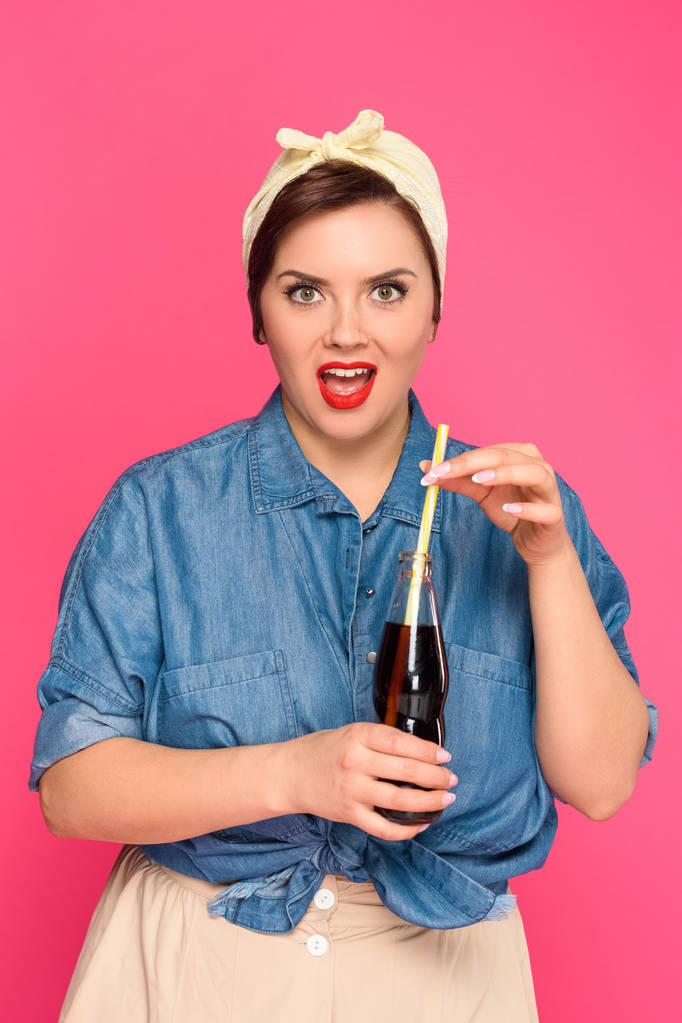 surprised size plus pin up woman holding glass bottle with drinking straw and looking at camera isolated on pink - Photo, Image