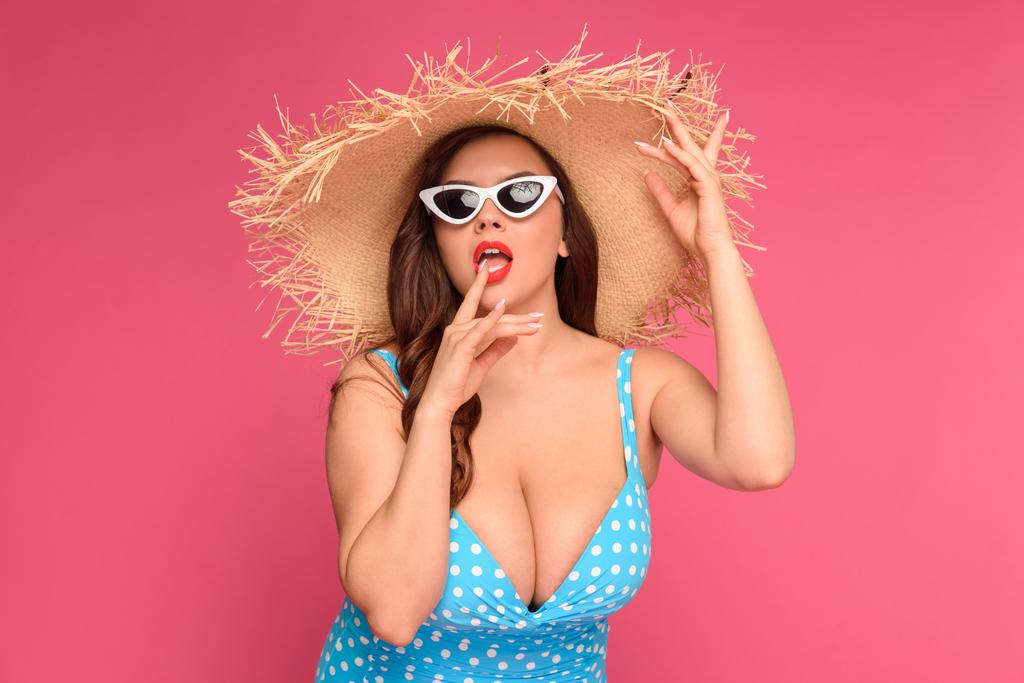 σέξι νεαρή μέγεθος συν γυναίκα με μαγιό φοράει ψάθινο καπέλο και γυαλιά ηλίου που απομονώνονται σε ροζ  - Φωτογραφία, εικόνα
