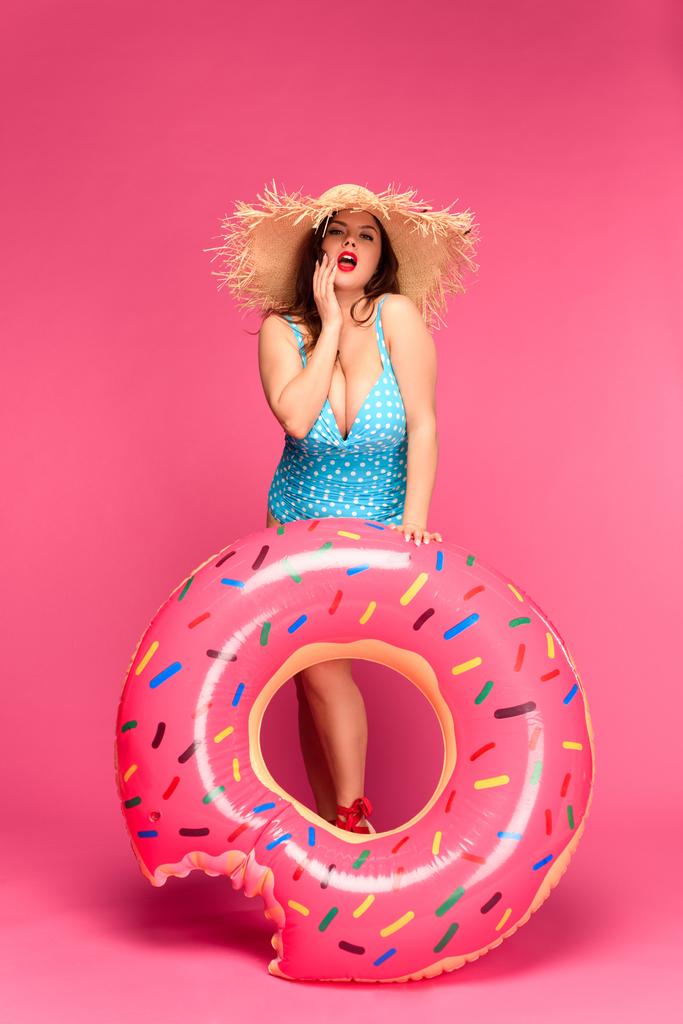 όμορφη μελαχρινή γυναίκα με μαγιό και άχυρο καπέλο κρατά φουσκωτό ντόνατ και βλέπουν τα φωτογραφικών μηχανών που απομονώνονται σε ροζ - Φωτογραφία, εικόνα