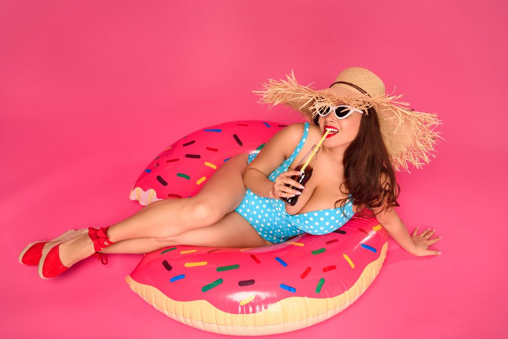 chica sexy en traje de baño y gafas de sol descansando en el anillo inflable y beber bebida refrescante aislado en rosa
 - Foto, Imagen