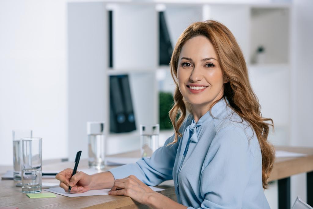side view of smiling businesswoman looking at camera at workplace with papers in office - Photo, Image