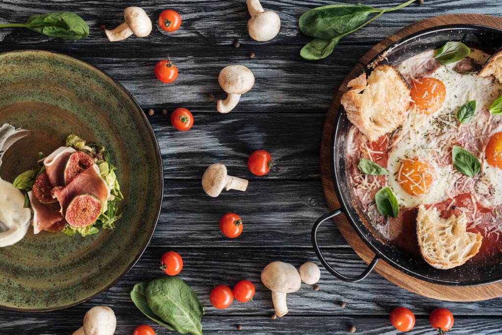 bovenaanzicht van heerlijke prosciutto, gebakken eieren en verse groenten op houten tafel - Foto, afbeelding