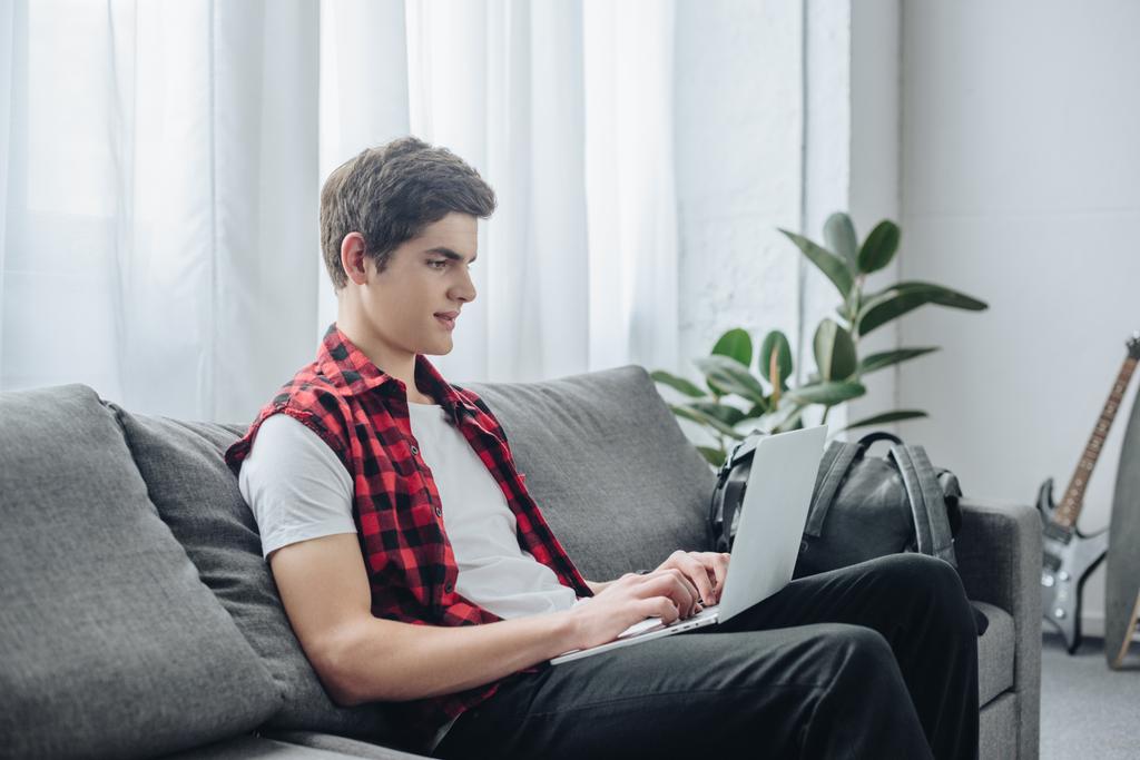 hübscher Teenager tippt auf Laptop, während er auf dem Sofa sitzt  - Foto, Bild