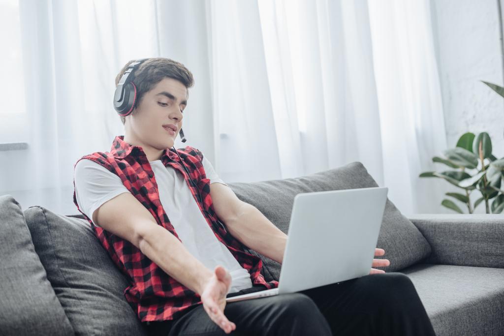 dospívající chlapec s sluchátka hrát hru na laptopu, zatímco sedí na pohovce  - Fotografie, Obrázek