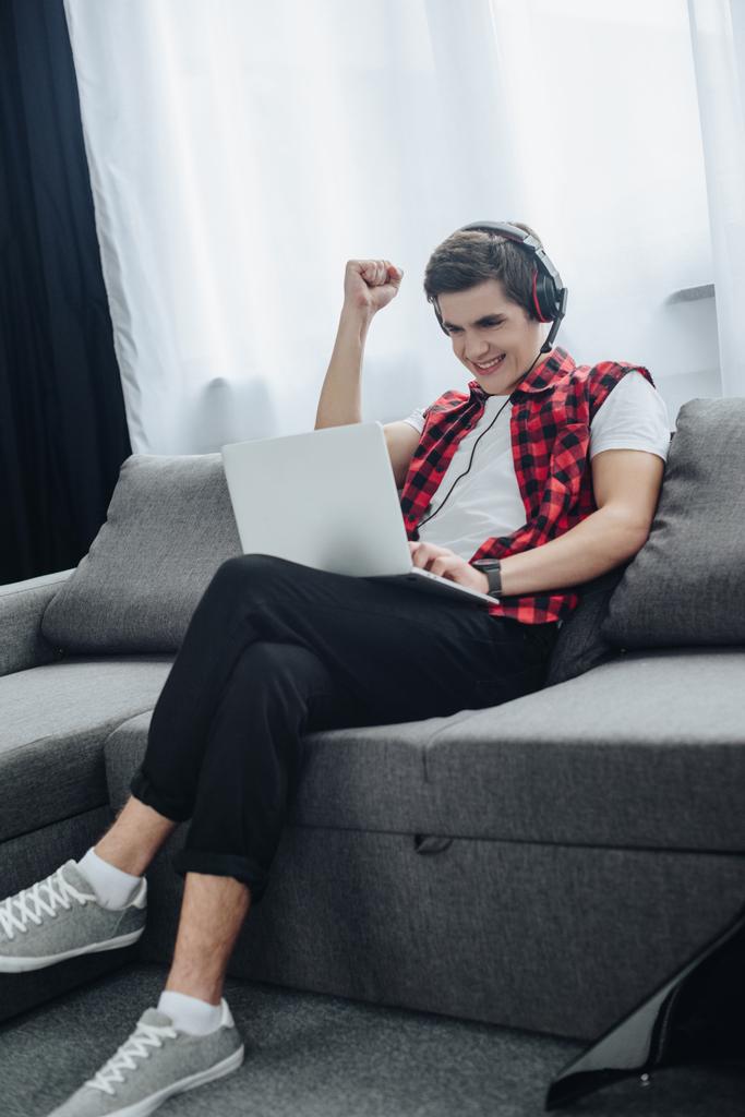 cheerful teenager with headphones playing game on laptop while sitting on sofa  - Photo, Image
