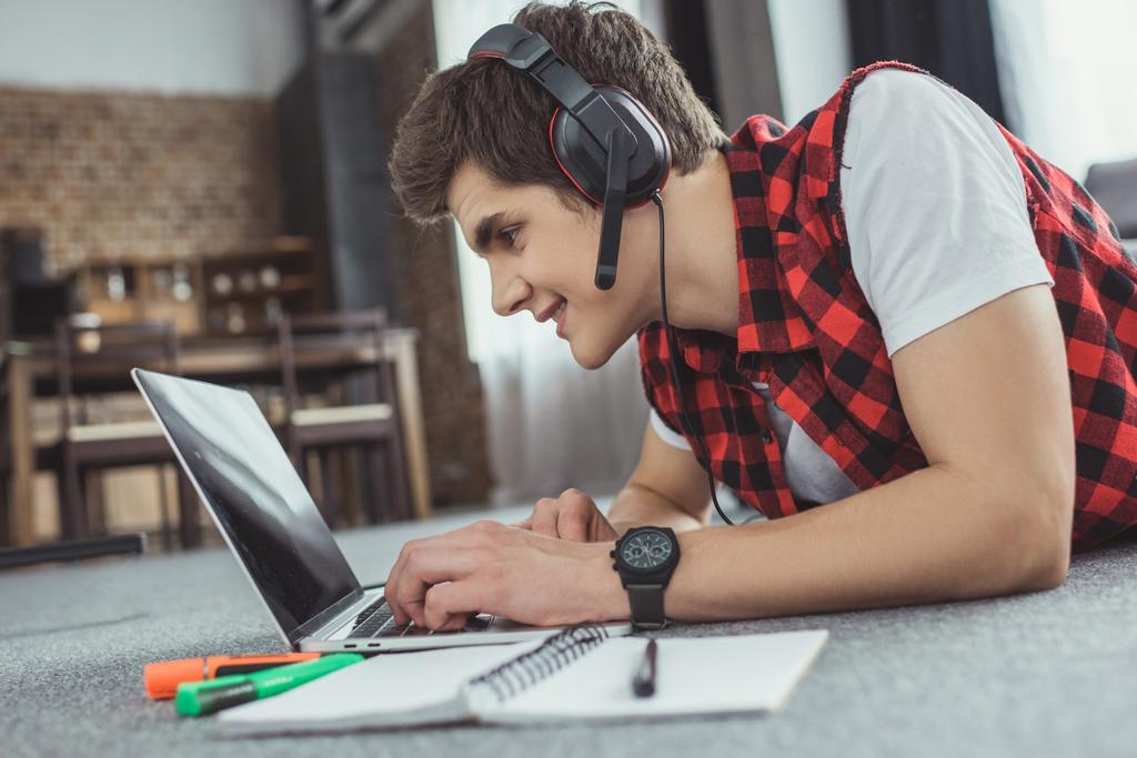 sonriente adolescente chico con auriculares jugando juego en el ordenador portátil mientras está acostado en el suelo con copybook
 - Foto, imagen