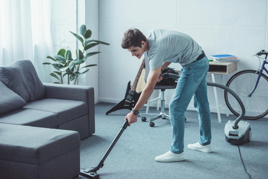 männlicher Teenager putzt Boden in der Nähe von Sofa mit Staubsauger - Foto, Bild