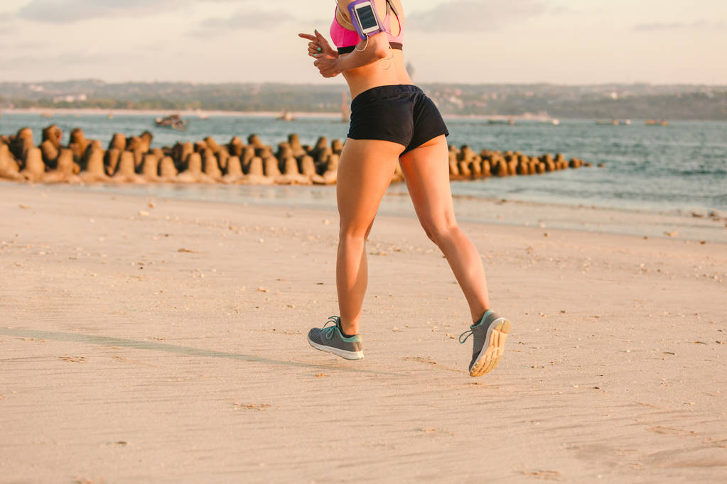 Oříznout záběr sportovkyně s smartphone v provozním náramkem případ běhání na pláži s mořem za - Fotografie, Obrázek