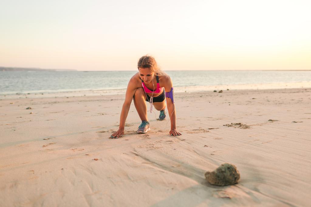 αθλήτρια στα ακουστικά με έξυπνο κινητό τηλέφωνο στην άσκηση σε αμμώδη παραλία με θάλασσα πίσω θήκη με περιβραχιόνιο - Φωτογραφία, εικόνα