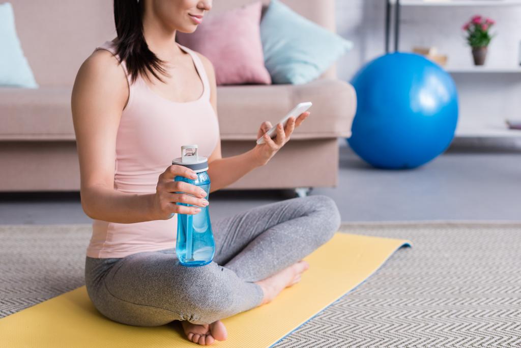 обрезанный снимок молодой женщины расслабляющей на циновке йоги после тренировки и использования смартфона дома
 - Фото, изображение