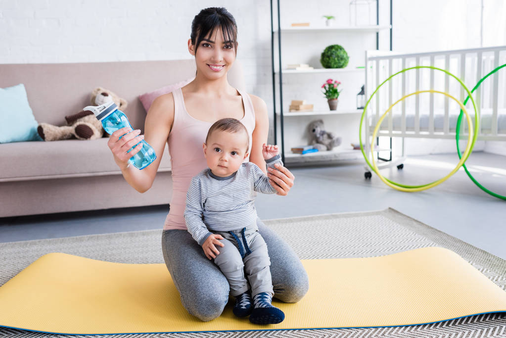 young beautiful mother and little child sitting on yoga mat at home and looking at camera - Photo, Image