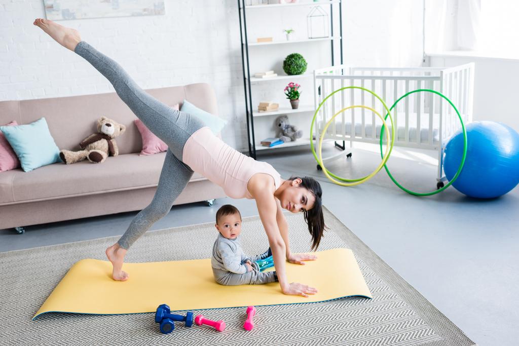 junge Mutter praktiziert Yoga in einbeiniger, nach unten gerichteter Hundehaltung, während ihr kleines Kind zu Hause auf Matte sitzt - Foto, Bild