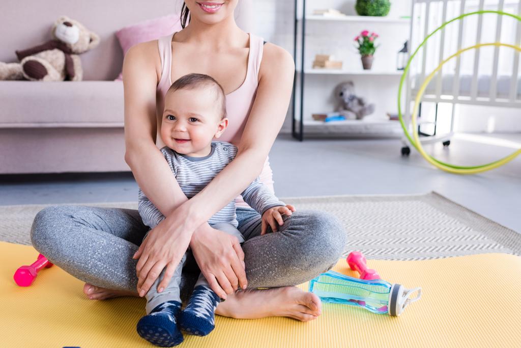 обрезанный снимок молодой спортивной матери и счастливого ребенка, сидящего на коврике для йоги дома
 - Фото, изображение