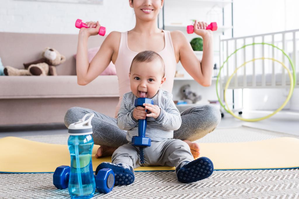 Schnappschuss einer lächelnden, fitten Mutter, die Übungen mit Kurzhanteln macht, während sie mit ihrem kleinen Kind zu Hause auf der Yogamatte sitzt - Foto, Bild