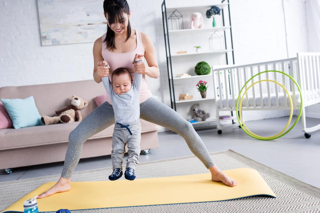 młoda matka sportowy, niosąc małe dziecko stojąc na matę do jogi w domu - Zdjęcie, obraz