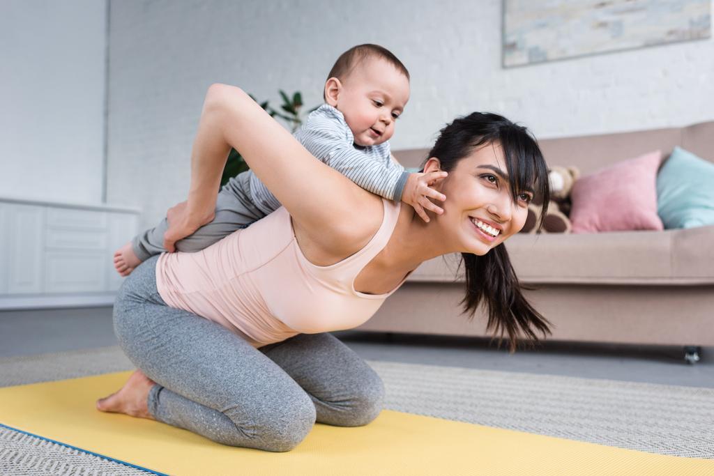 Маленький ребенок свисает на мать, пока она стоит на коврике для йоги дома
 - Фото, изображение