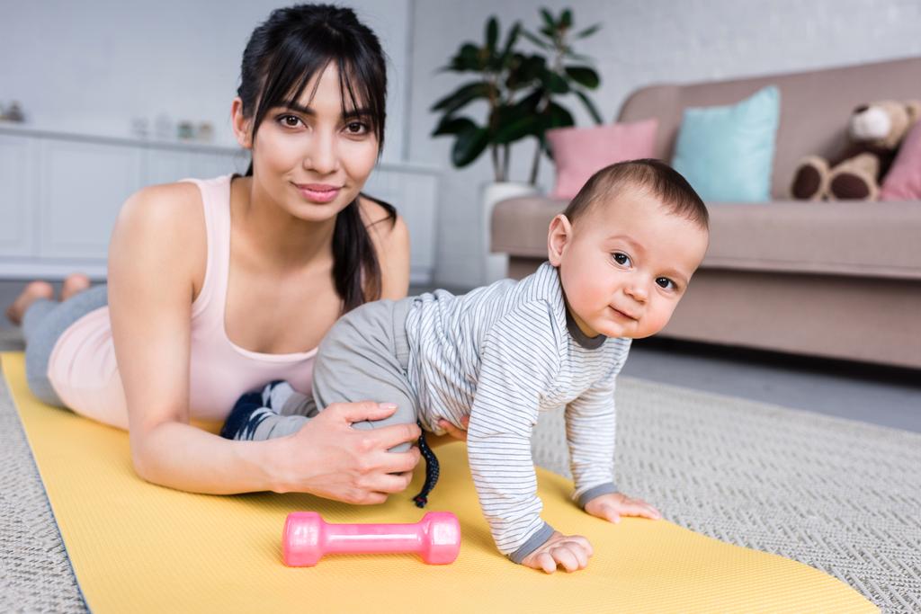 junge glückliche Mutter und kleines Kind zu Hause auf Yogamatte und schauen in die Kamera - Foto, Bild